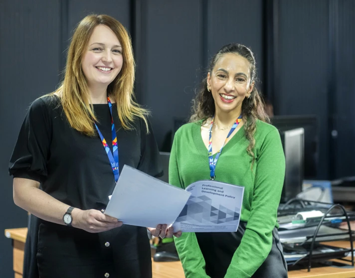 Two Glasgow Kelvin College support staff members smiling and holding documents in an office environment, with a colleague working at a desk in the background. Two Glasgow Kelvin College support staff members smiling and holding documents in an office environment, with a colleague working at a desk in the background.